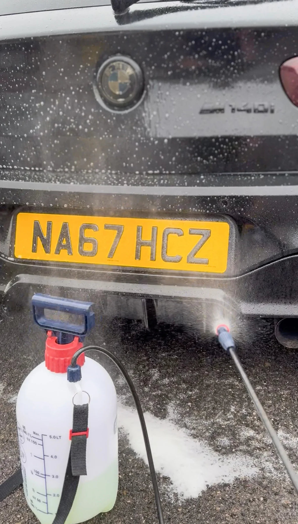 Pre-wash foam being applied to a black BMW rear bumper with a pump sprayer during car detailing
