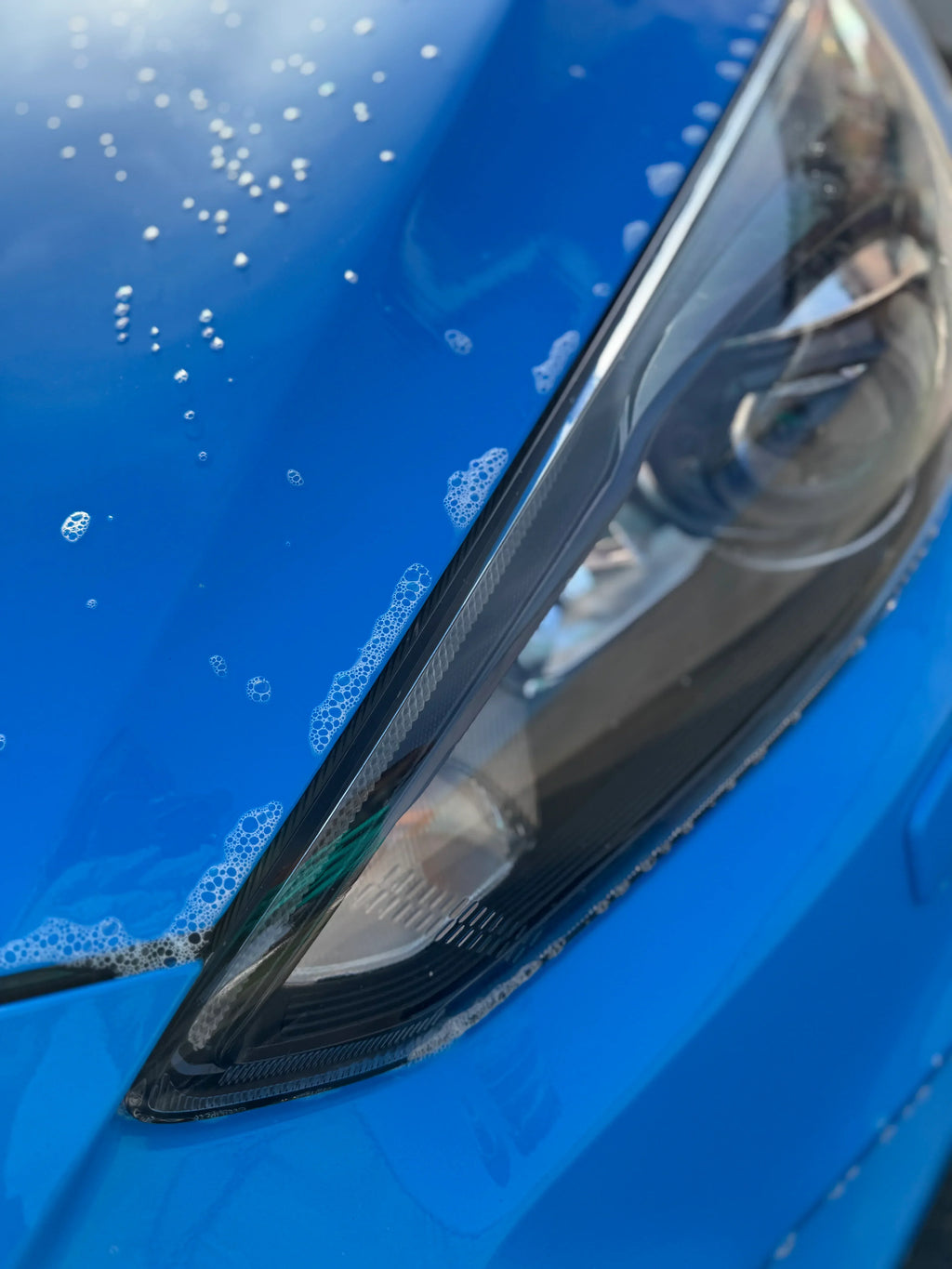 Close-up of blue car hood and headlight with soap suds, car detailing and cleaning